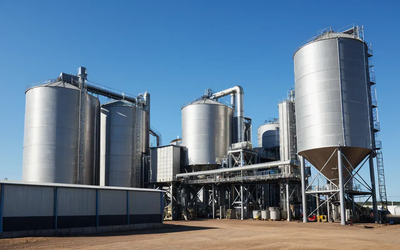 A modern ready-mix batching plant with large storage silos and mixing drums under a clear blue sky with industrial piping visible in the foreground.